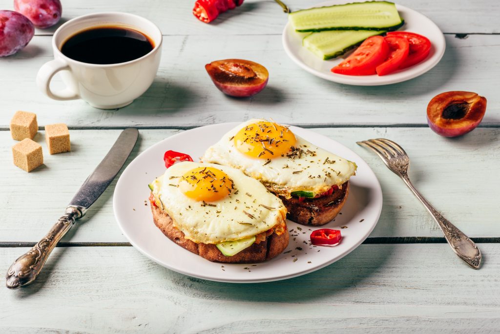 Bruschettas with vegetables and fried egg on white plate, cup of coffee and some fruits over wooden background. Healthy food concept.